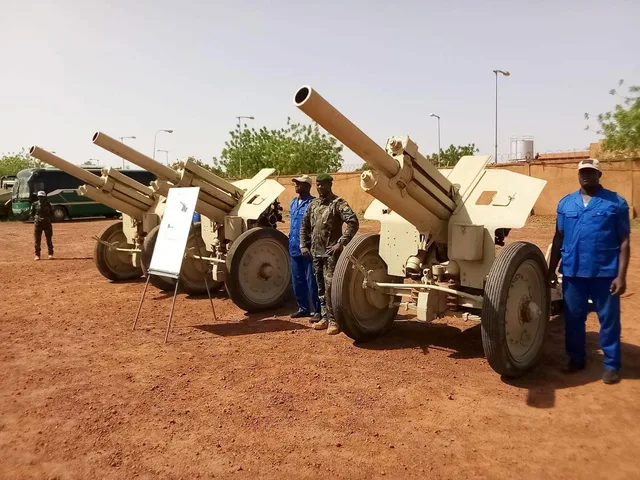 ثلاثة مدافع ميدانية على أرض ترابية حمراء يقف بجانبها عسكري وعمال بزي أزرق، صورة من الأرشيف