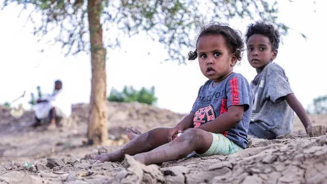 طفلان سودانيان صغيران يجلسان على أرض جافة متشققة تحت شجرة، وفي الخلفية رجل جالس، صورة أرشيفية
