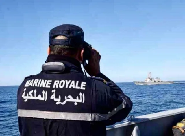 عنصر من البحرية الملكية المغربية يرتدي سترة كتب عليها اسمها يراقب البحر بمنظار وفي الأفق سفينة، صورة أرشيفية