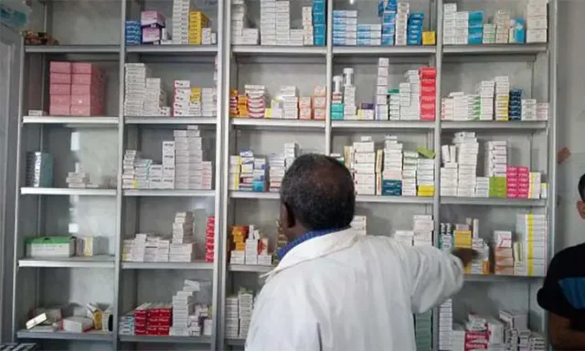 صيدلي بمعطف أبيض يظهر من الخلف وهو يتناول علبة من رفوف مليئة بالأدوية، صورة توضيحية أرشيفية